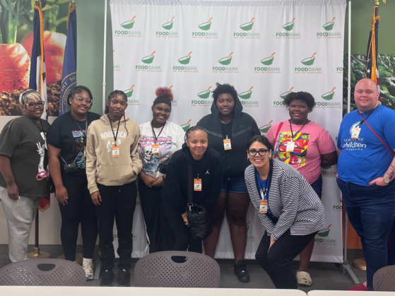 A diverse group of teens pose in front of a Food Bank for the Heartland backdrop with Voices for Children staff